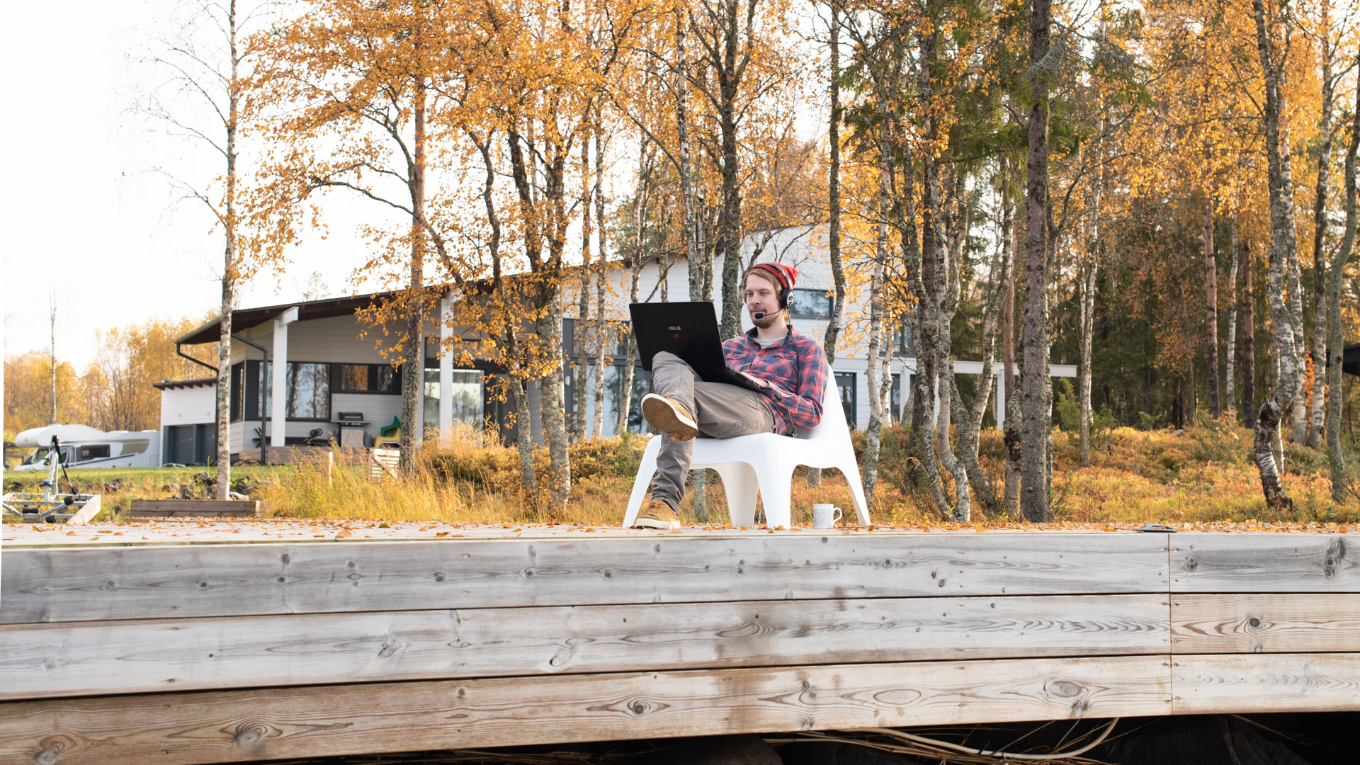 Hipsterimies tekee töitä ruska-aikaan annalla tietokoneella. Hänen päässään on kuulokemikrofoni. Taustalla on omakotitalo.