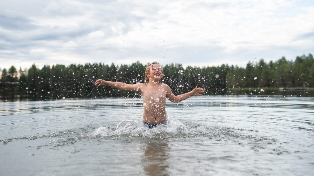 Poika kahlaa ja nauraa Helinlammen rantavedessä ja vesi pärsyy.