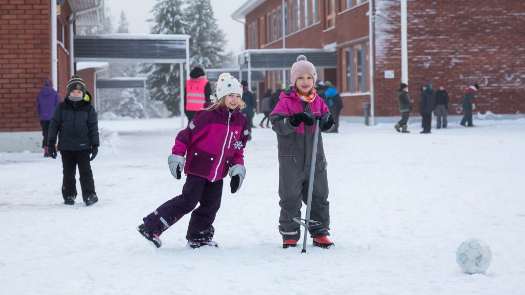 Rukan koulun oppilaat potkaisevat palloa ja hyppivät hymyillen koulun lumisella pihalla.