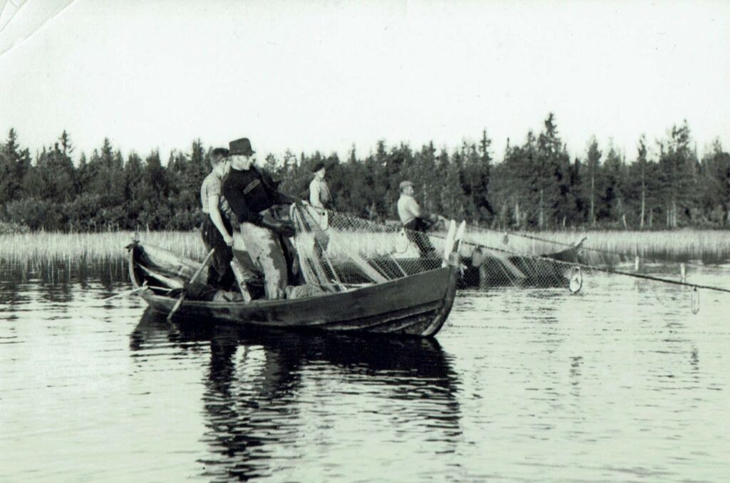 Mustavalkoinen kuva, jossa kalastajia seisoo puuveneessä ja vetää verkkoja vedestä.