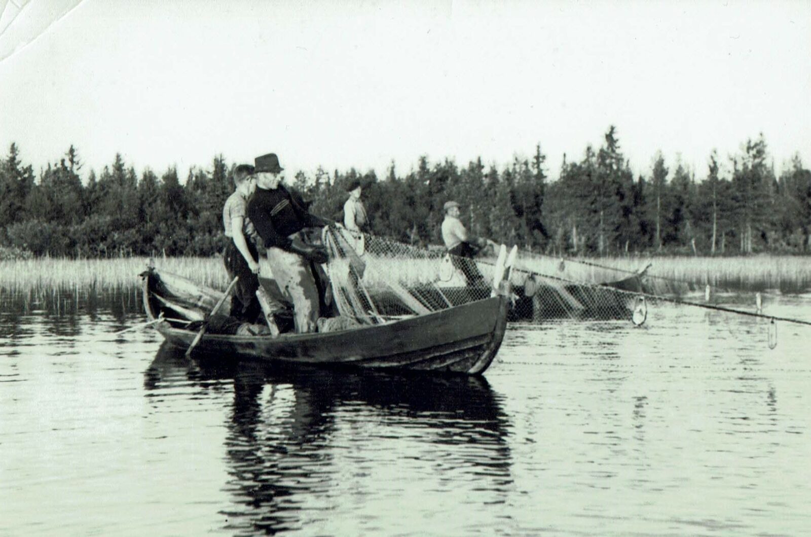 Mustavalkoinen kuva, jossa kalastajia seisoo puuveneessä ja vetää verkkoja vedestä.